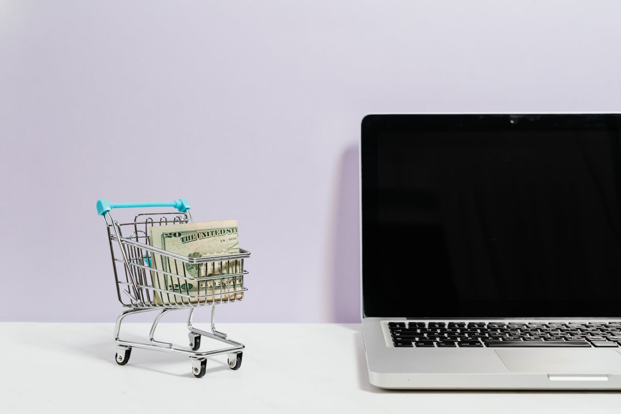 a mini shopping cart with money inside sits next to an open laptop with a black screen.